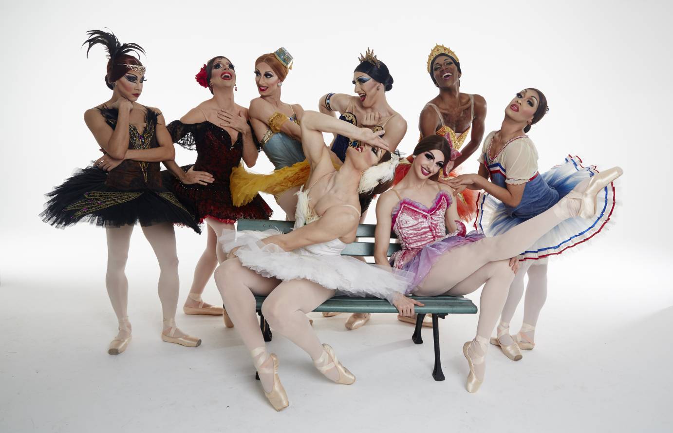 Wearing colorful classical tutus and heavy make up, the dancers pose with silly faces. Two dancers sit on a bench while six stand behind them.